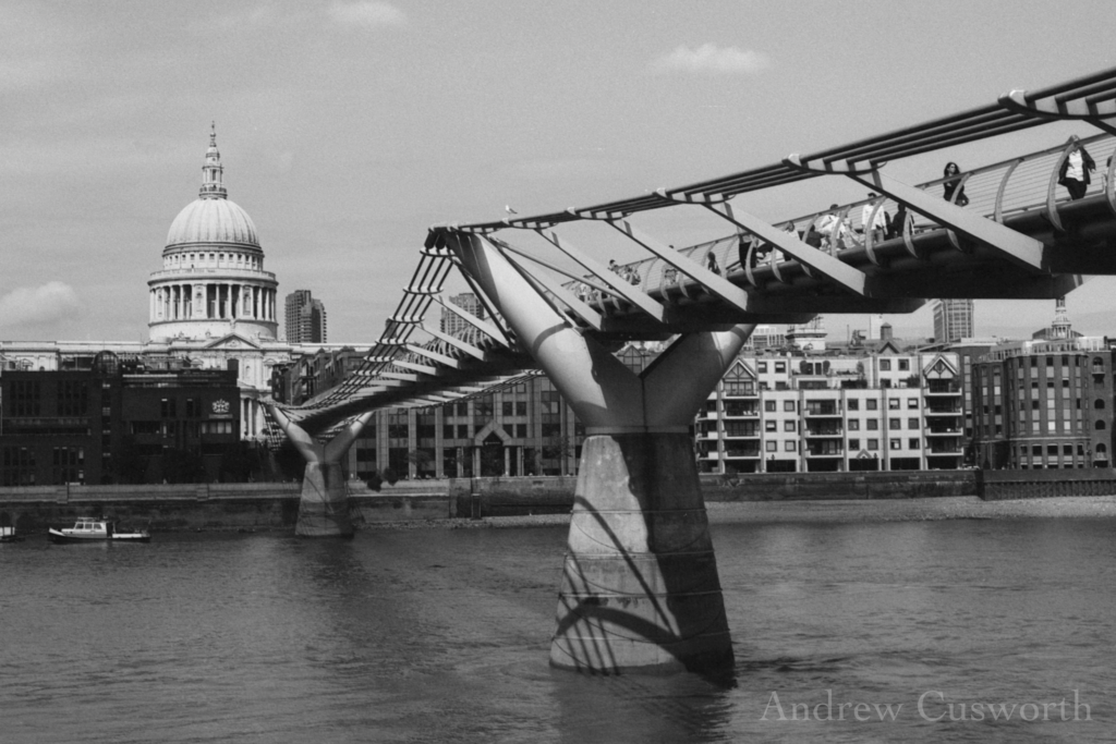A black and white photograph showing St Paul's Cathedral and the Millennium Bridge seen from the South side of the Thames. The sky is clear, and bright sun falls on the bridge and the cathedral.