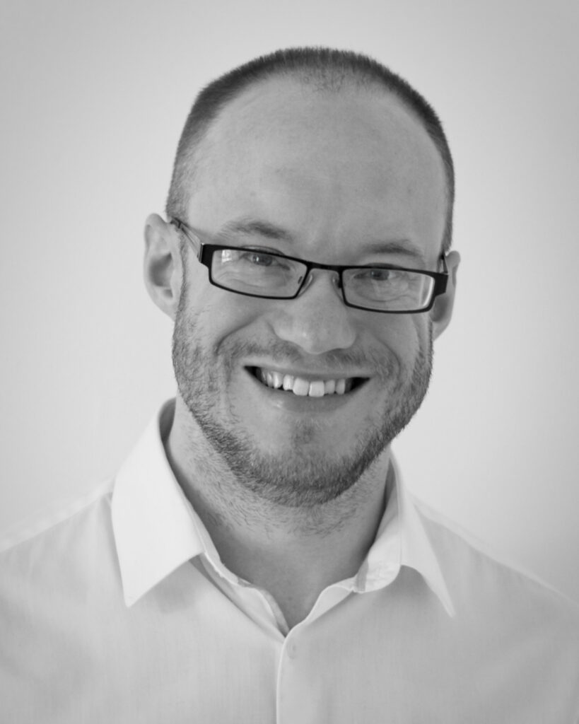 A black and white photograph of Andrew Cusworth wearing a white shirt and glasses smiling into the lens.